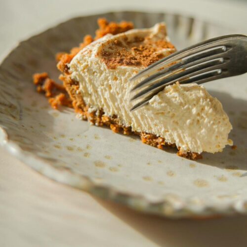 A slice of creamy pumpkin cheesecake with a crumbly graham cracker crust rests on a textured plate. A fork is cutting into the cake, casting a soft shadow across the plate. The scene is warmly lit, highlighting the cake's smooth texture.